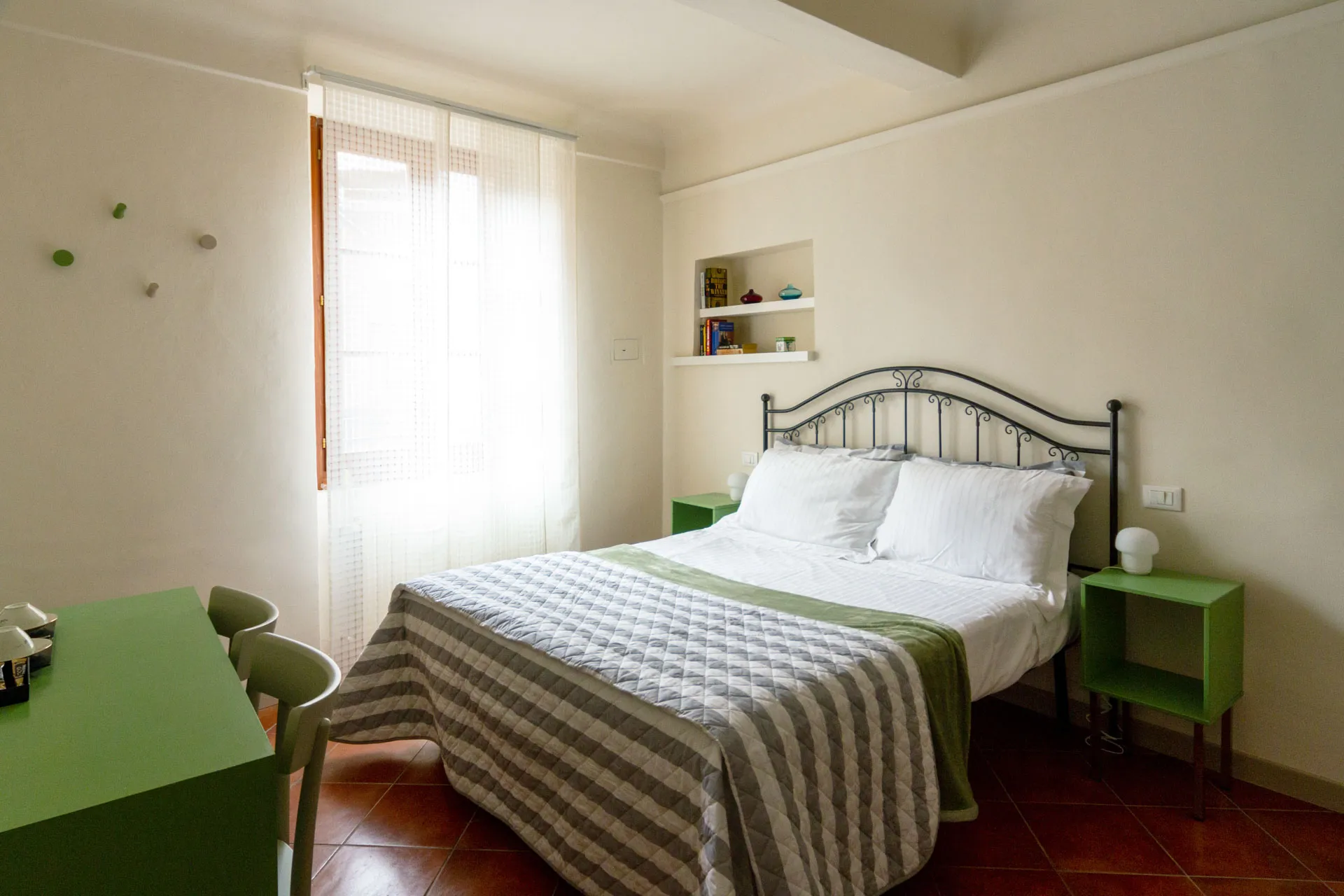 Double bedroom at B&B San Francesco in Siena, featuring a wrought iron bed, striped grey and white quilt, green nightstands, and terracotta flooring. A large window with white curtains fills the room with natural light.