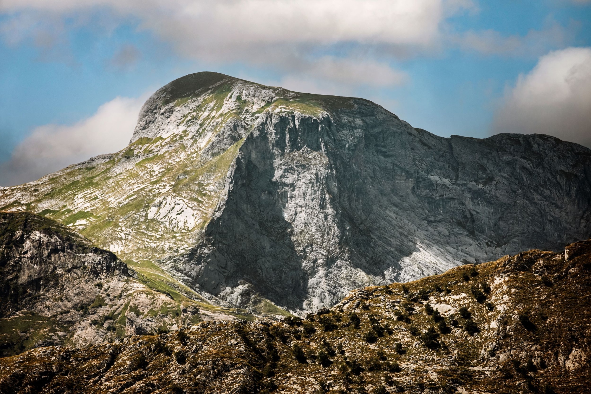 Maestoso picco roccioso delle Alpi Apuane con pendii ripidi di roccia bianca e grigia, prati verdi in primo piano e cielo azzurro