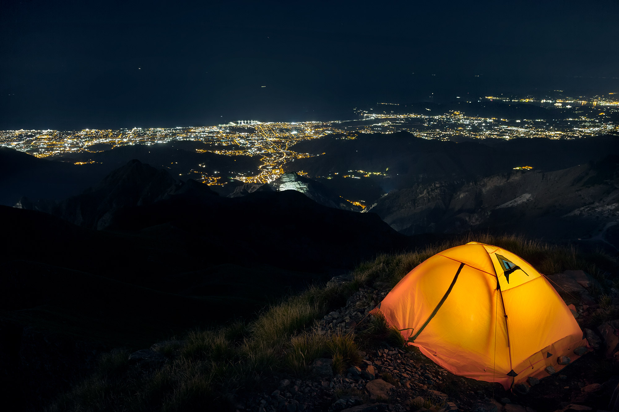 Tenda da campeggio gialla illuminata su un pendio erboso del Monte Corchia di notte, con vista panoramica sulle luci delle città della costa toscana e sulle Alpi Apuane sullo sfondo