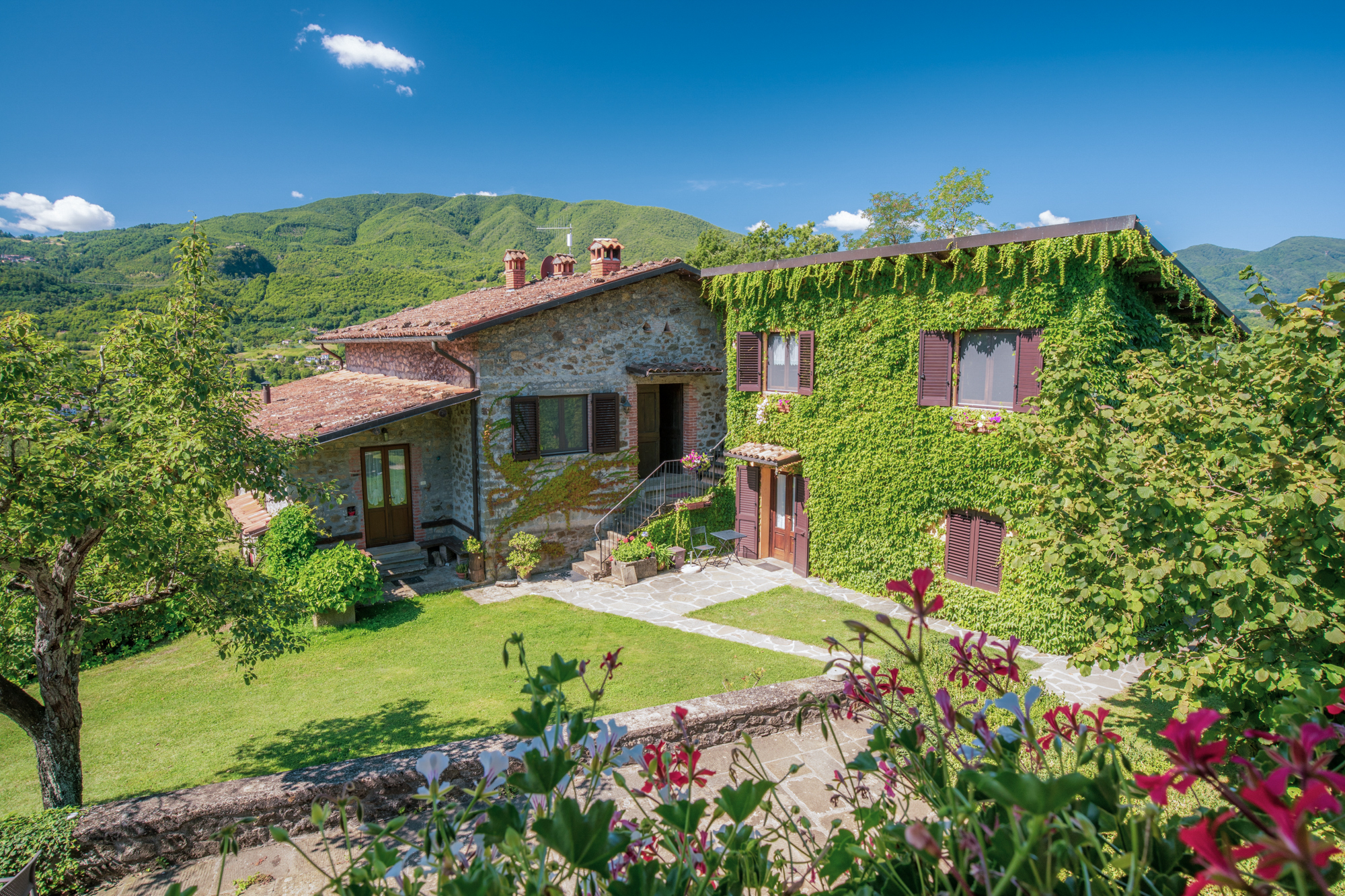 Agriturismo Braccicorti circondato da giardino fiorito con margherite rosse e vegetazione rigogliosa, edificio in pietra con muri chiari con finestre in legno scuro e edera verde che copre parzialmente la struttura
