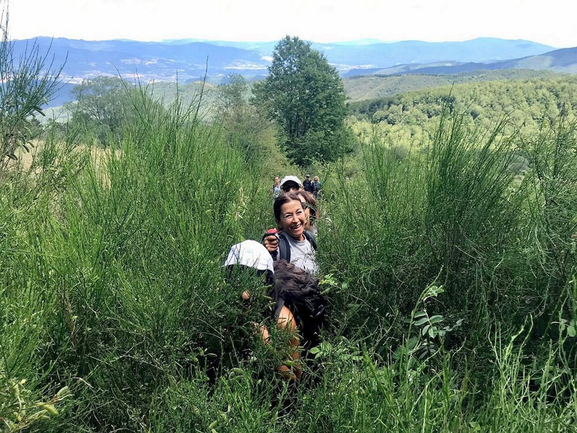 Gruppo di escursionisti che percorre un sentiero immerso nella vegetazione alta dell'Appennino Pratese, con panorama sulle colline boscose del Mugello sullo sfondo. Una donna sorridente in primo piano tiene un bastoncino da trekking.