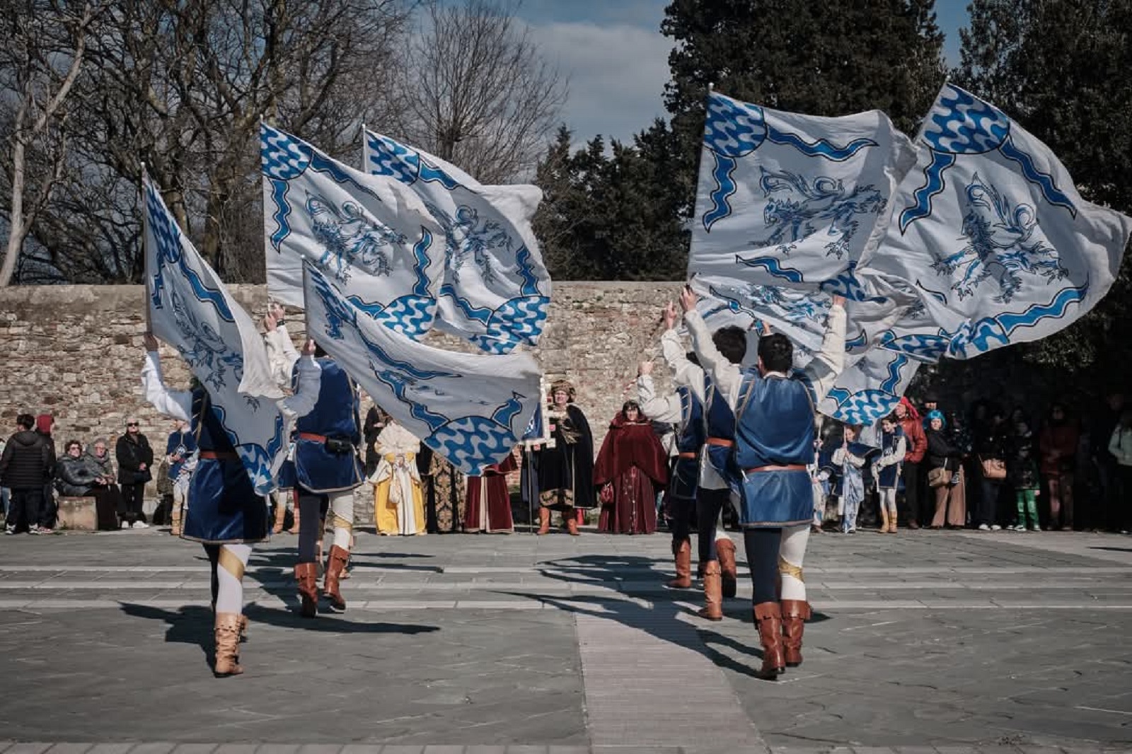 Sbandieratori in costume storico si esibiscono con bandiere bianche e azzurre in una piazza, davanti al pubblico e ad altri figuranti.
