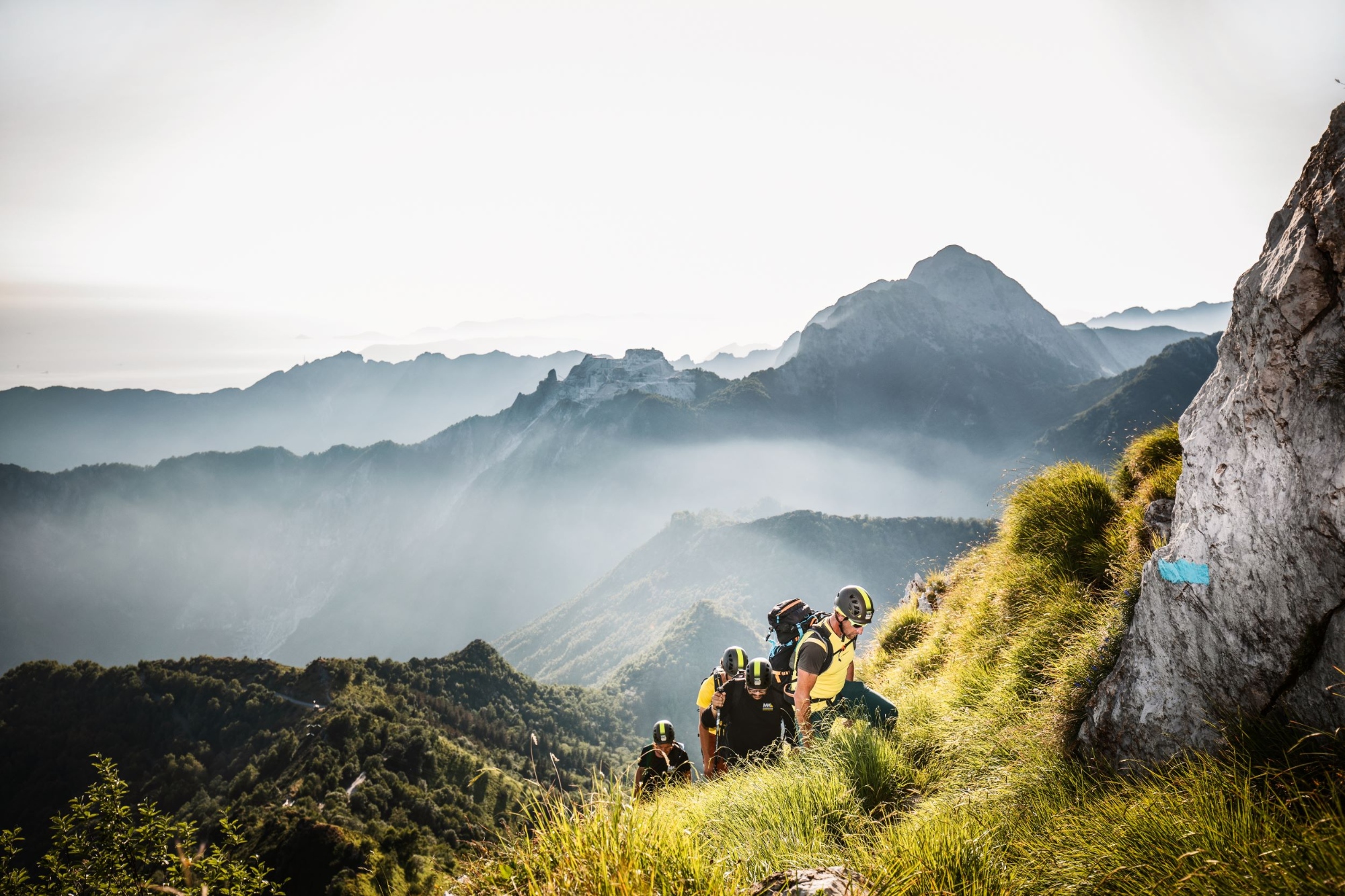 Escursionisti con zaini gialli e neri percorrono un sentiero di montagna circondati da pareti rocciose, con una vista della valle nebbiosa e le montagne delle Apuane sullo sfondo