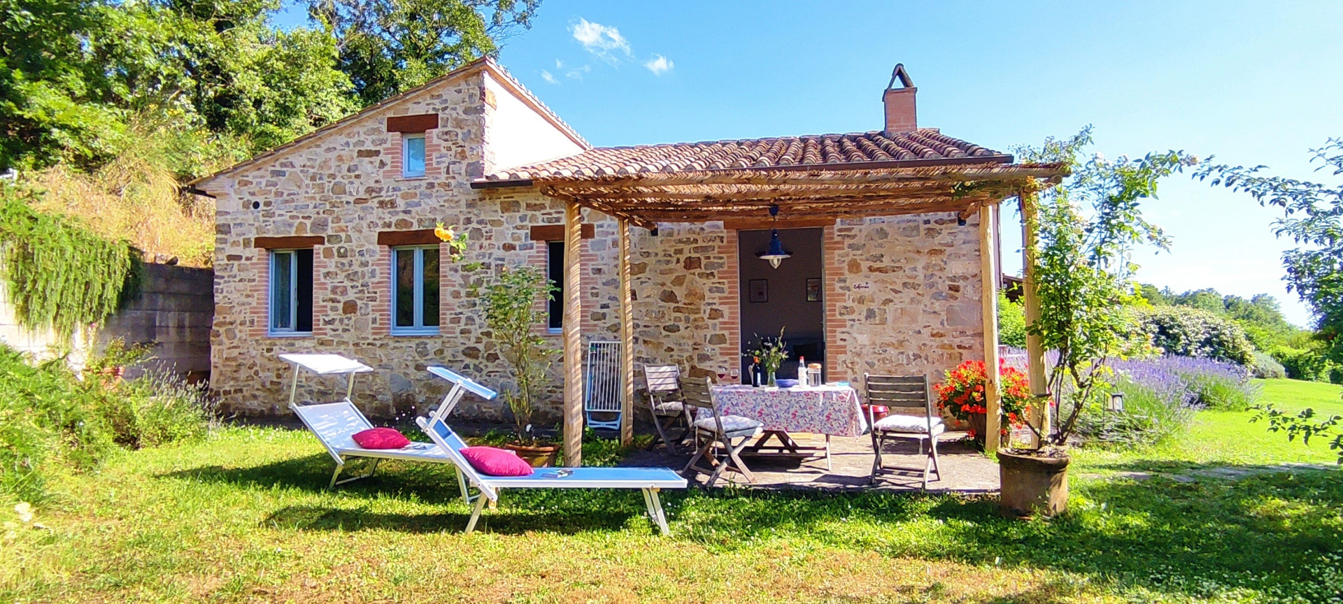 Esterno di un casale in pietra tipico toscano con un grande giardino verde. Davanti alla struttura si trova un patio coperto da un pergolato in legno con tavolo e sedie, mentre sul prato ci sono due lettini prendisole. Sullo sfondo si notano alberi e piante di lavanda in fiore sotto un cielo azzurro.