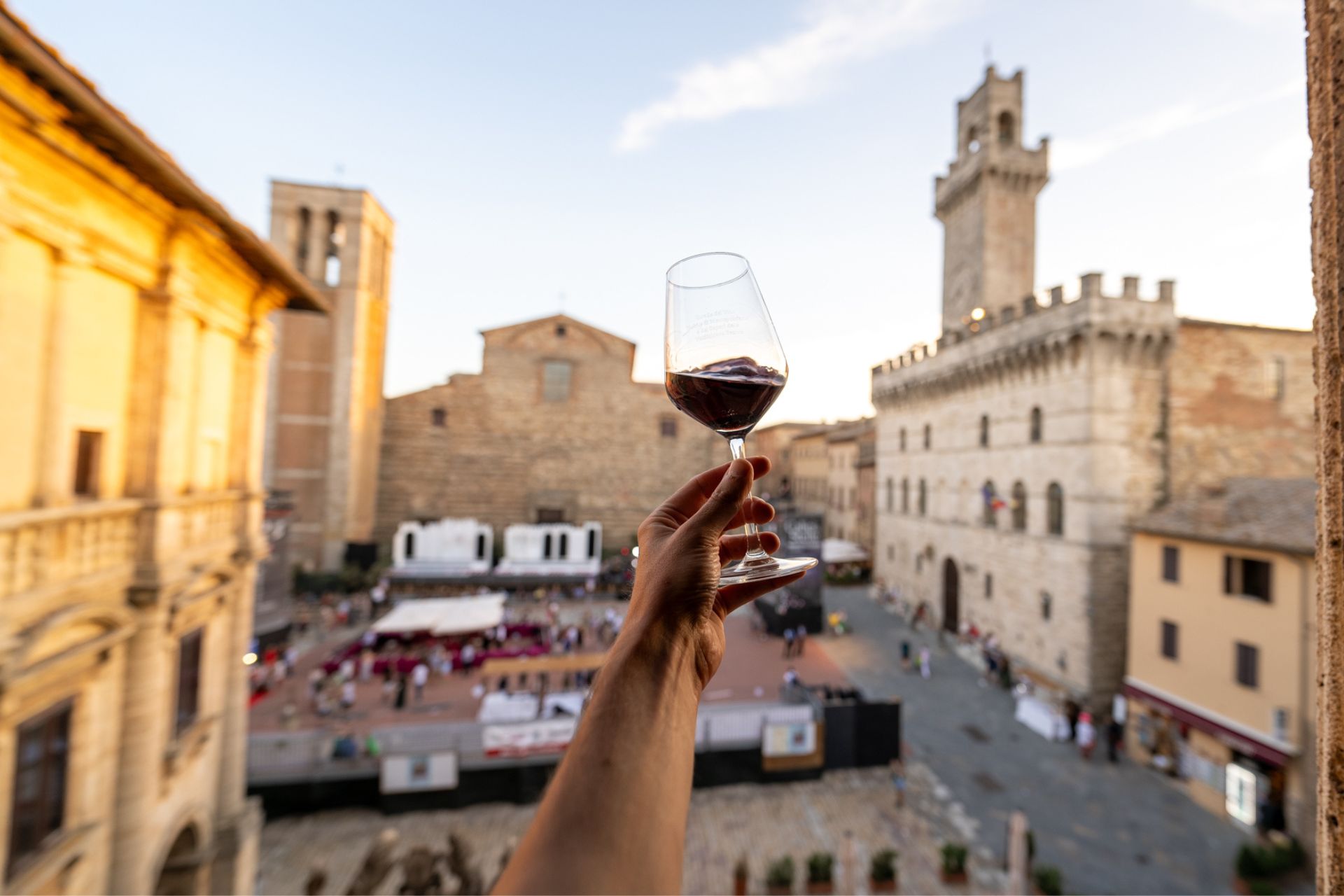 Eine Hand hebt ein Glas Rotwein in den Vordergrund. Im Hintergrund ist die Piazza Grande von Montepulciano mit ihren historischen Gebäuden im Licht des Sonnenuntergangs zu sehen. Menschen schlendern über den Platz während einer Weinveranstaltung