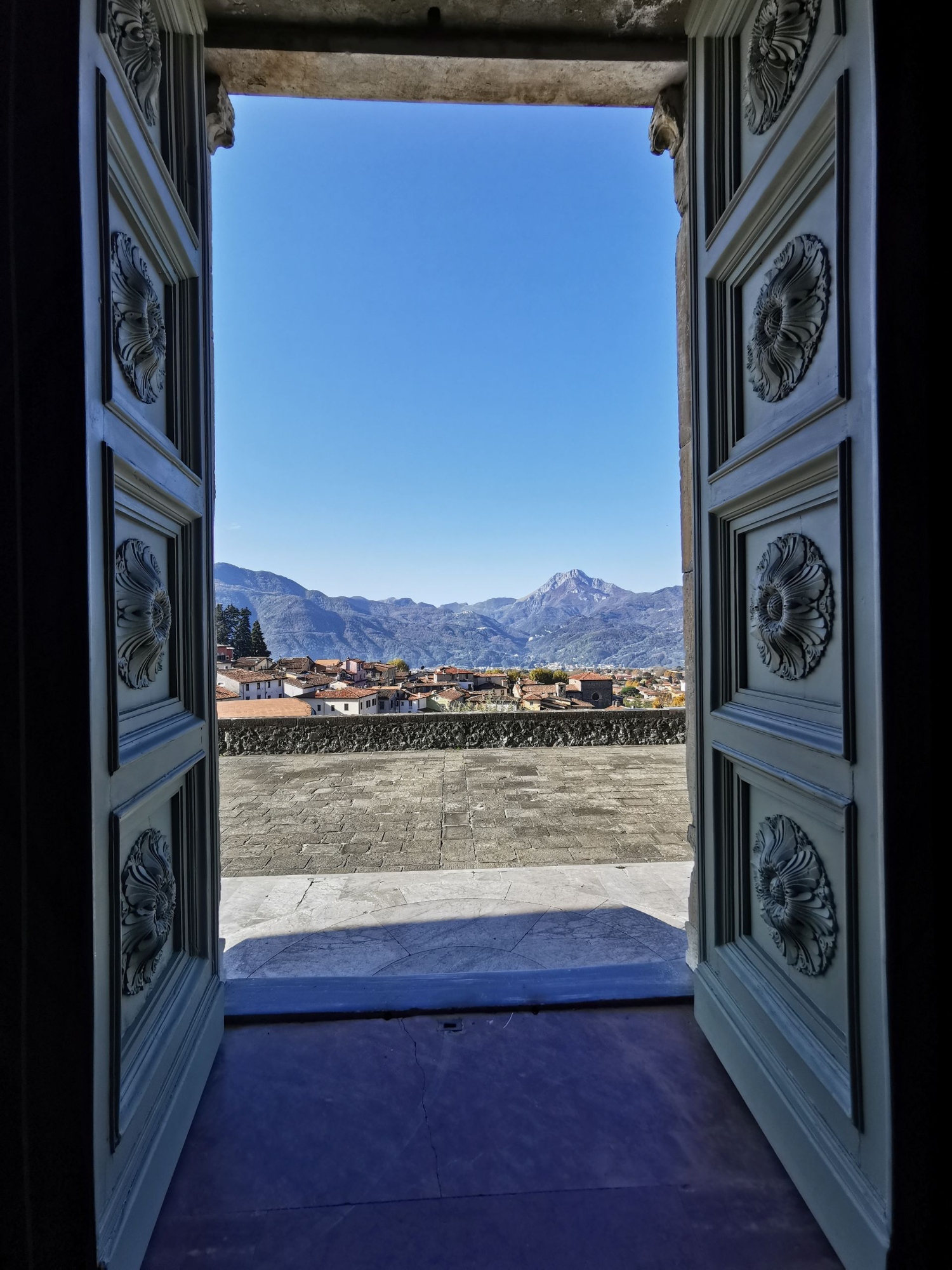 Vista incorniciata da un elegante portone decorato a rilievo floreale aperto su una terrazza panoramica di Barga, con i tetti del borgo medievale e le Alpi Apuane sullo sfondo sotto un cielo azzurro