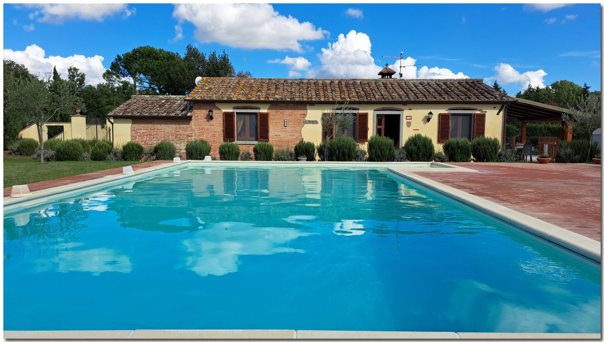 A low-angle perspective view looking across the private swimming pool of Villino Cortona, a traditional Tuscan brick and stucco cottage. The turquoise water perfectly reflects a bright blue sky with white, fluffy clouds. In the background stands the cottage with dark wooden shutters, a manicured hedge of boxwood and lavender, and olive trees. To the right, the terracotta-tiled sun deck features several potted plants in terracotta urns. The scene is sun-drenched and relaxing.