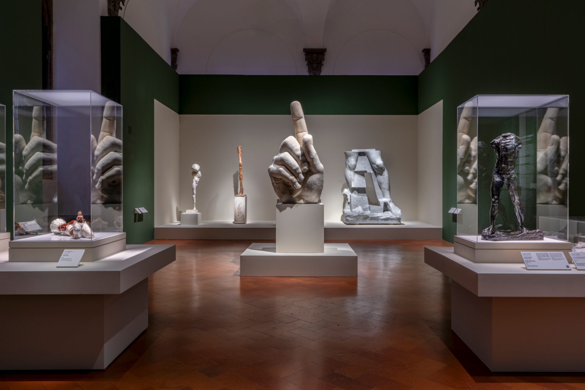 One of the rooms of Palazzo Strozzi with sculptures by Auguste Rodin displayed in cases, including a bronze figure. At the center, a large hand with its index finger raised.