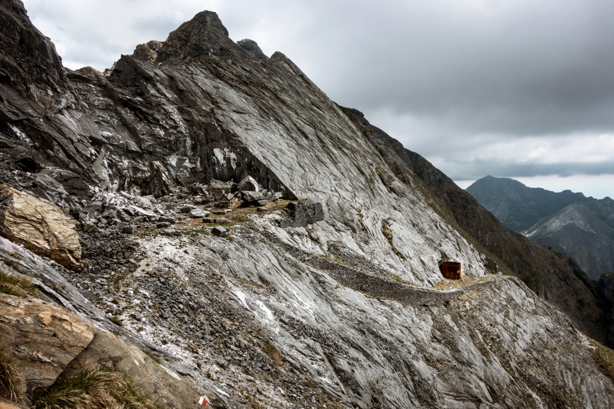 Cava di marmo apuana con struttura di legno rosso (baracca) sulle rocce bianche e grigie stratificate, circondata da montagne boscose sullo sfondo sotto cielo nuvoloso