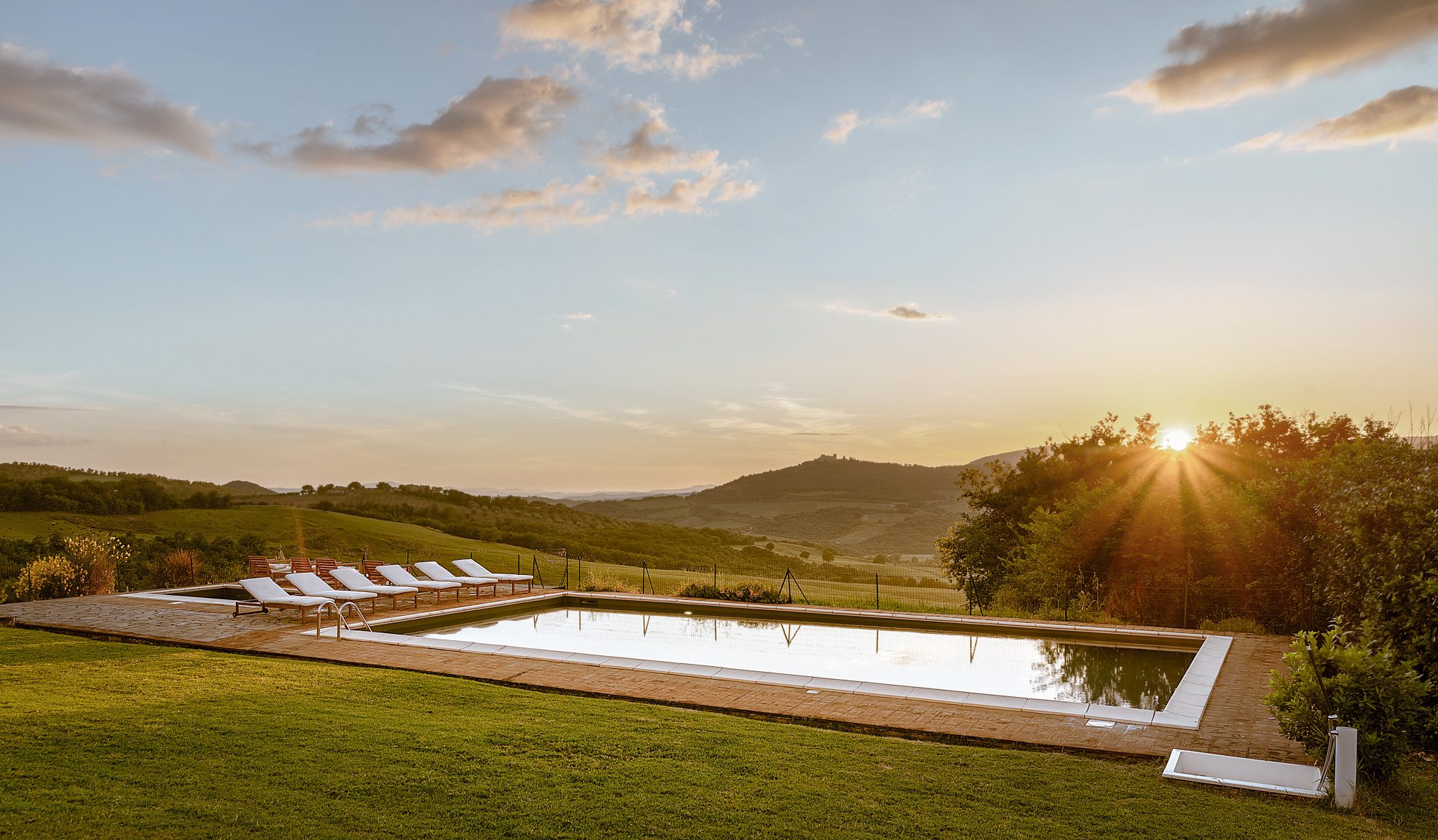 Piscina panoramica all'aperto di un agriturismo in Toscana circondata da prato verde e colline al tramonto con lettini prendisole.