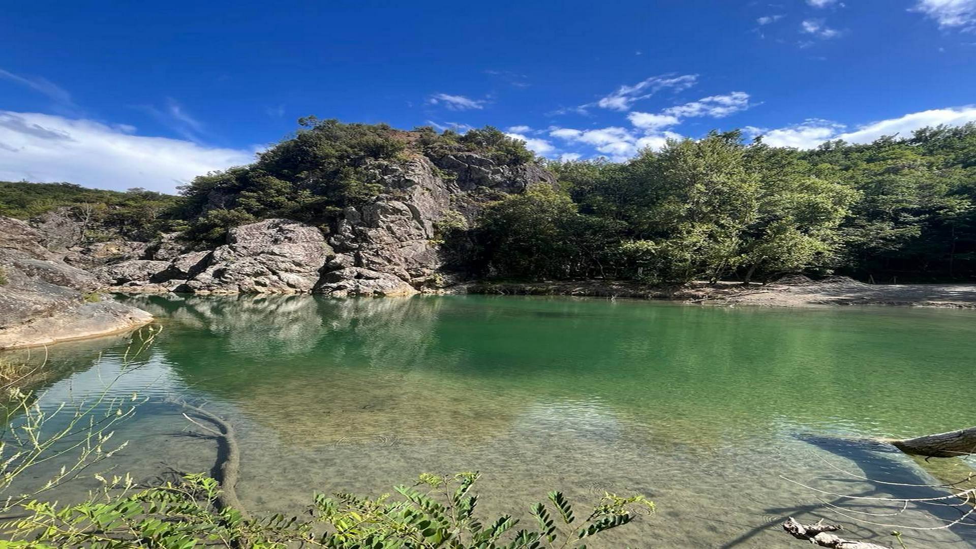 Pozza naturale del fiume Merse con acqua verde-smeraldo cristallina ai piedi di una parete rocciosa grigia; vegetazione mediterranea incornicia la riva in una giornata di sole con cielo blu