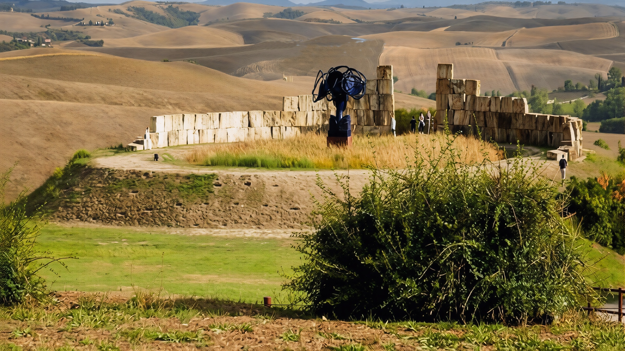 Curved travertine block walls of the Teatro del Silenzio in Lajatico with a dark metal sculpture at the centre and visitors walking along the path, surrounded by the bare rolling hills of the Valdera