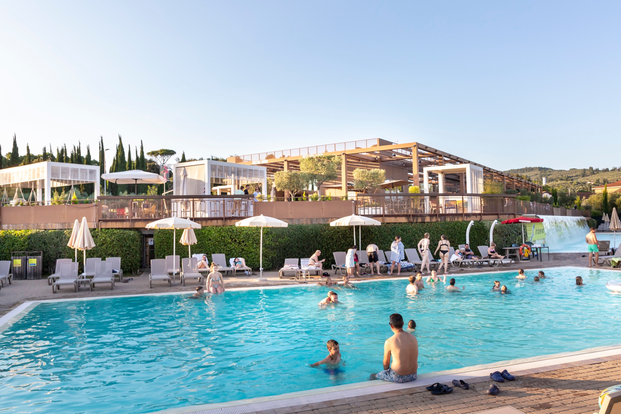 Genitori e bambini in piscina con alle spalle la struttura centrale del campeggio con tettoie