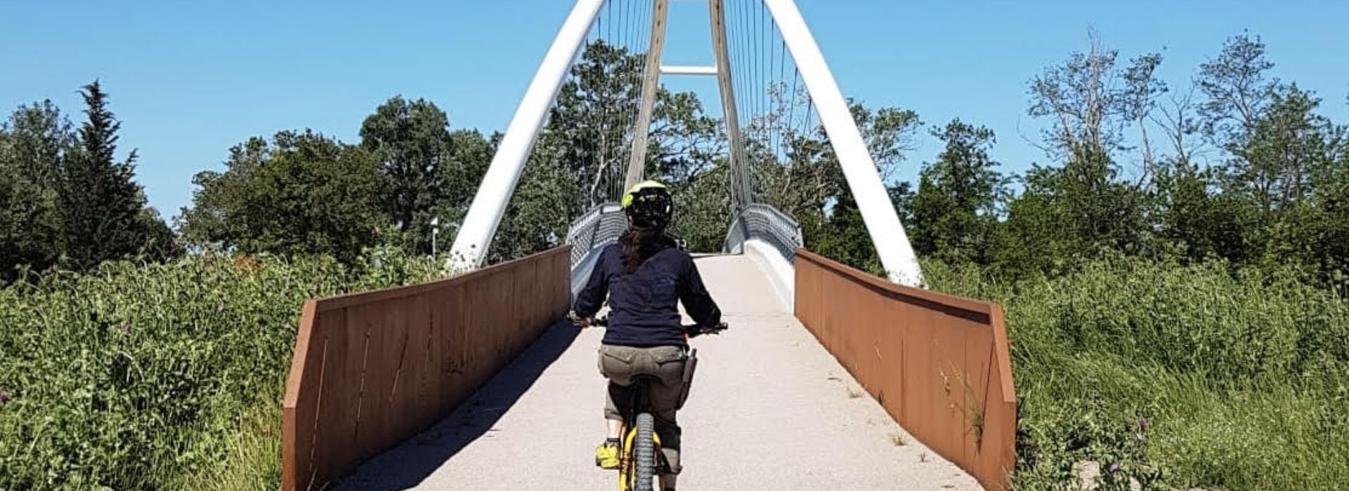 Ciclista in e-bike con casco giallo attraversa un ponte ciclopedonale ad arco bianco immerso nella natura della Maremma toscana, tra vegetazione rigogliosa e cielo azzurro