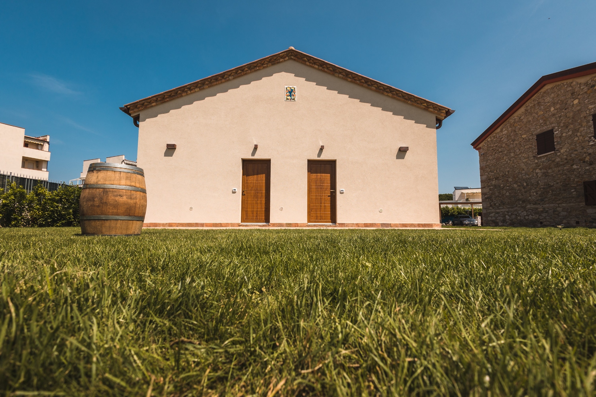 Inquadratura dal basso del prato verde curato di una struttura ricettiva o cantina in stile toscano. Sullo sfondo si staglia la facciata simmetrica di un edificio intonacato di chiaro, caratterizzato da due porte d'ingresso indipendenti in legno scuro e un tetto a spiovente. Sulla sinistra del prato è posizionata una grande botte in legno decorativa, mentre sulla destra si intravede la parete in pietra di una struttura adiacente, il tutto sotto un cielo estivo azzurro e limpido.