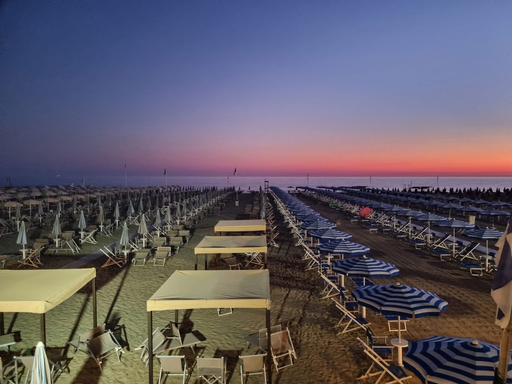 Spiaggia attrezzata del Bagno Venezia a Lido di Camaiore al tramonto, con file di ombrelloni bianchi e blu a righe, lettini e gazebo sulla sabbia, sullo sfondo il mare del Tirreno con il cielo che sfuma dal viola all'arancio rosso all'orizzonte