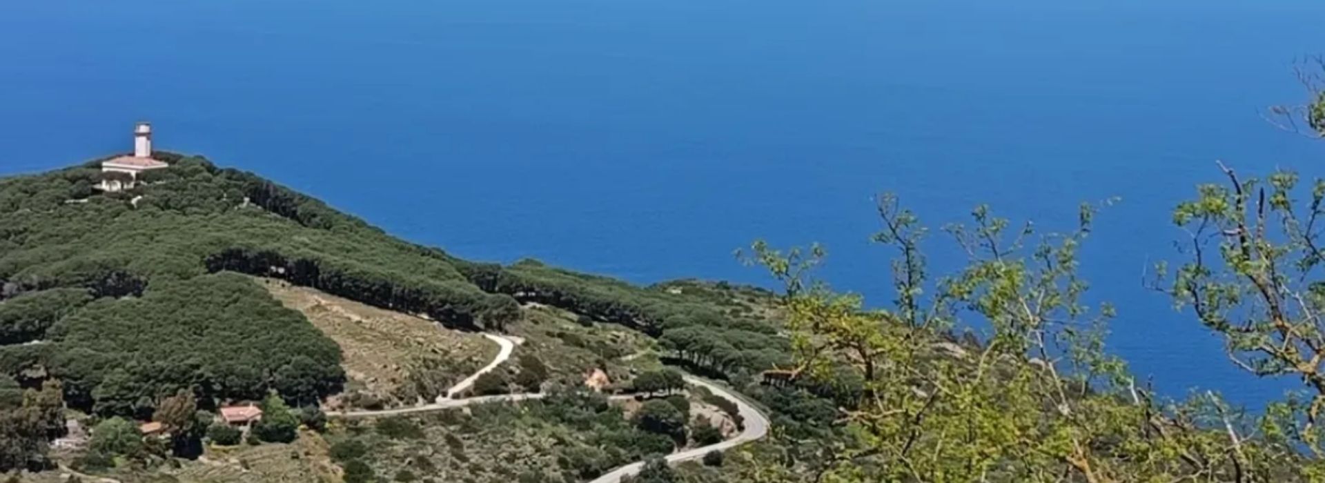 Dirt road winding through the Mediterranean scrub towards the Capel Rosso lighthouse, atop a hill on Giglio Island, with the azure sea of ​​the Tuscan Archipelago in the background