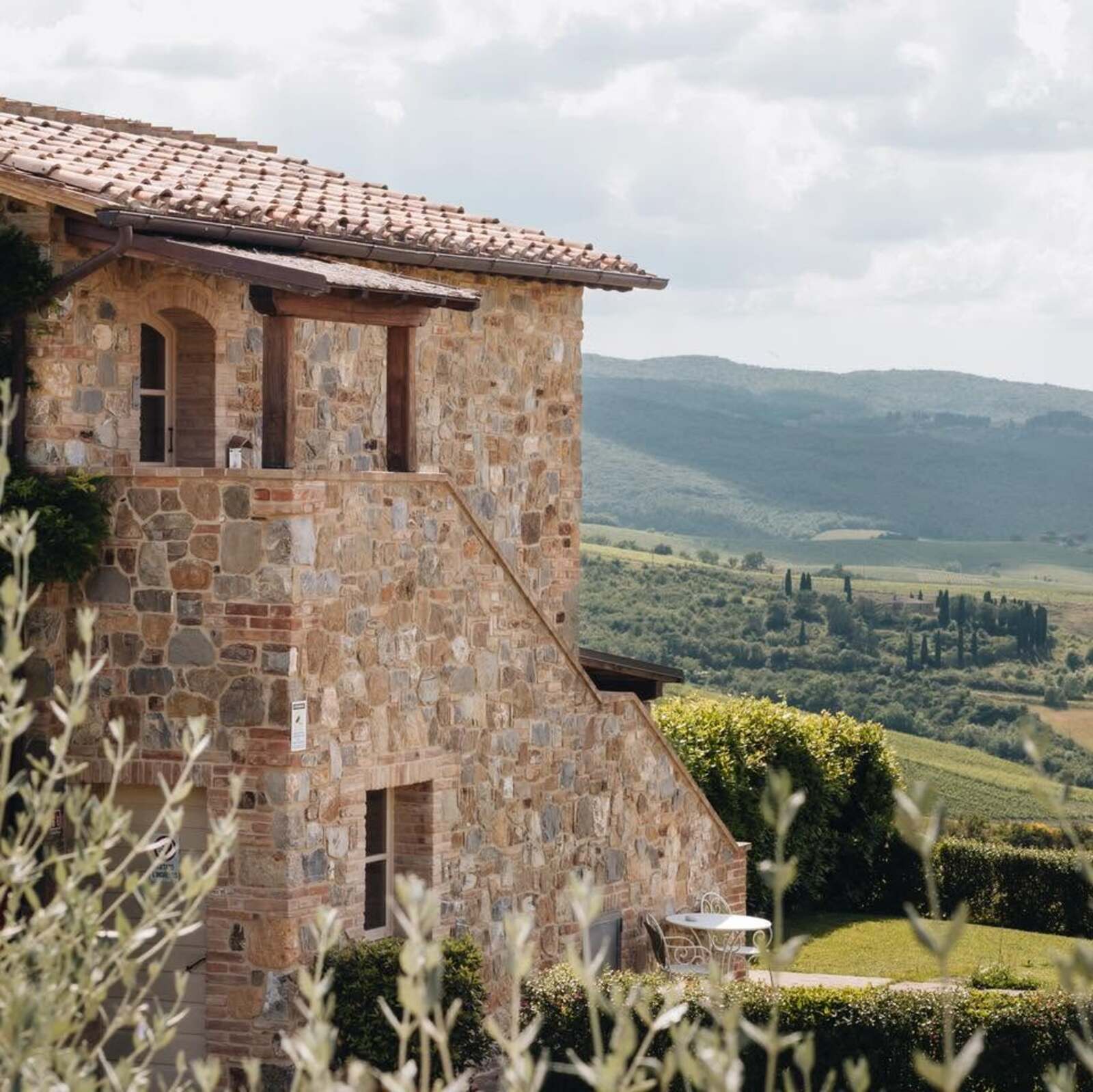 Close-up of a traditional Tuscan stone farmhouse featuring a terracotta tile roof and external stone stairs. The upper balcony shows rustic wooden beams and an arched window. Below, a manicured garden features a small white wrought-iron table and chairs surrounded by green hedges. In the background, a breathtaking panoramic view of the Tuscan countryside unfolds with rolling green hills, vineyards, and iconic cypress trees under a partly cloudy sky.