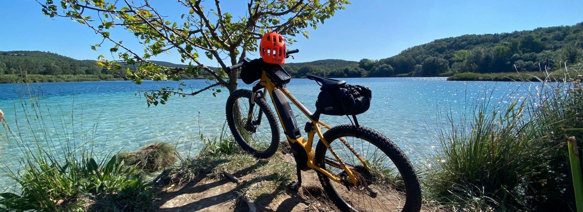 E-bike gialla con casco rosso appoggiata a un albero sulla riva del Lago dell'Accesa, con acque cristalline turchesi e colline boscose sullo sfondo, durante un tour in bicicletta nelle Colline Metallifere in Maremma