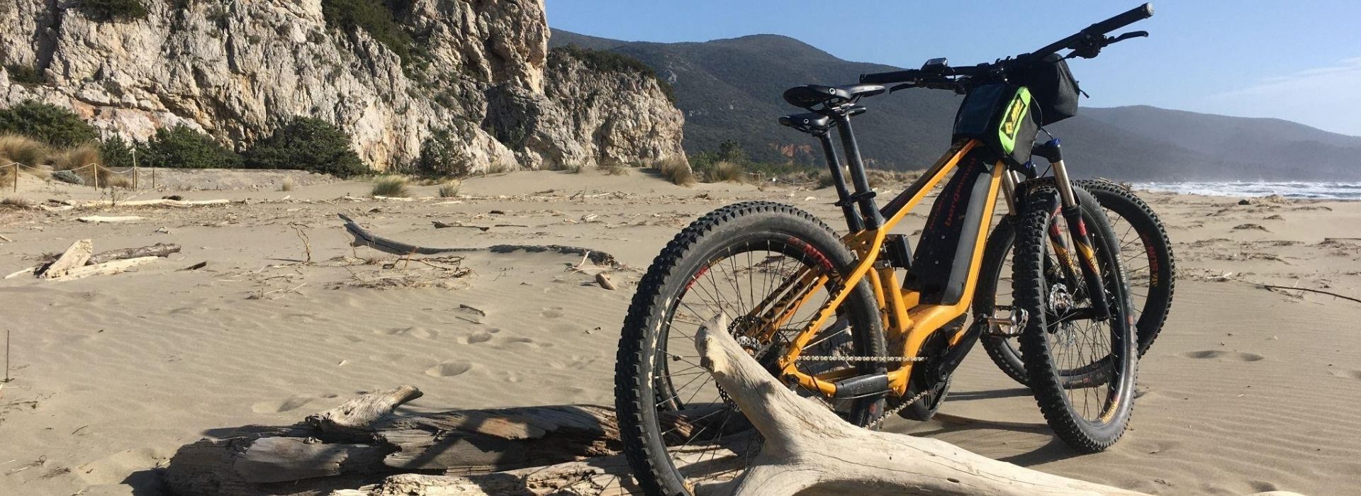 Two orange e-bikes parked on a driftwood log on Collelungo Beach, in the Maremma Natural Park, with rocky cliffs and wooded hills in the background