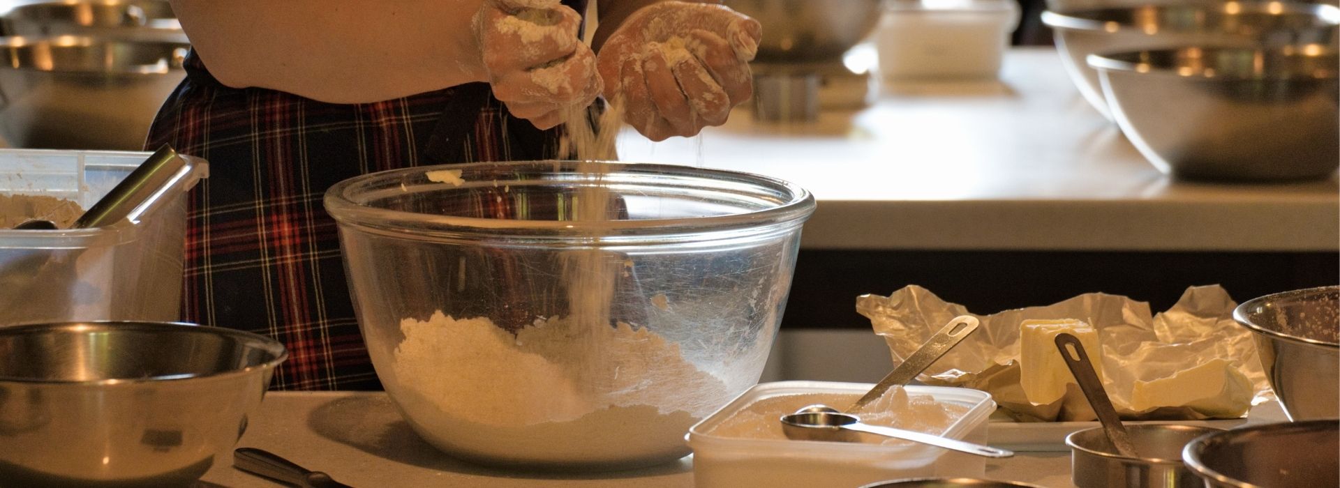 Floured hands sift flour into a glass bowl during a cooking class in Maremma, with butter, utensils, and steel bowls on the worktop