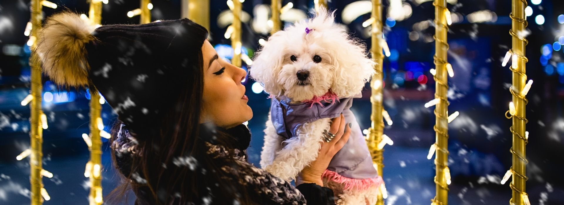 Woman in fur-lined winter hat holding a Maltese dog dressed in a coat, amidst twinkling golden lights and falling snow, during a pet-friendly New Year's Eve holiday in Tuscany