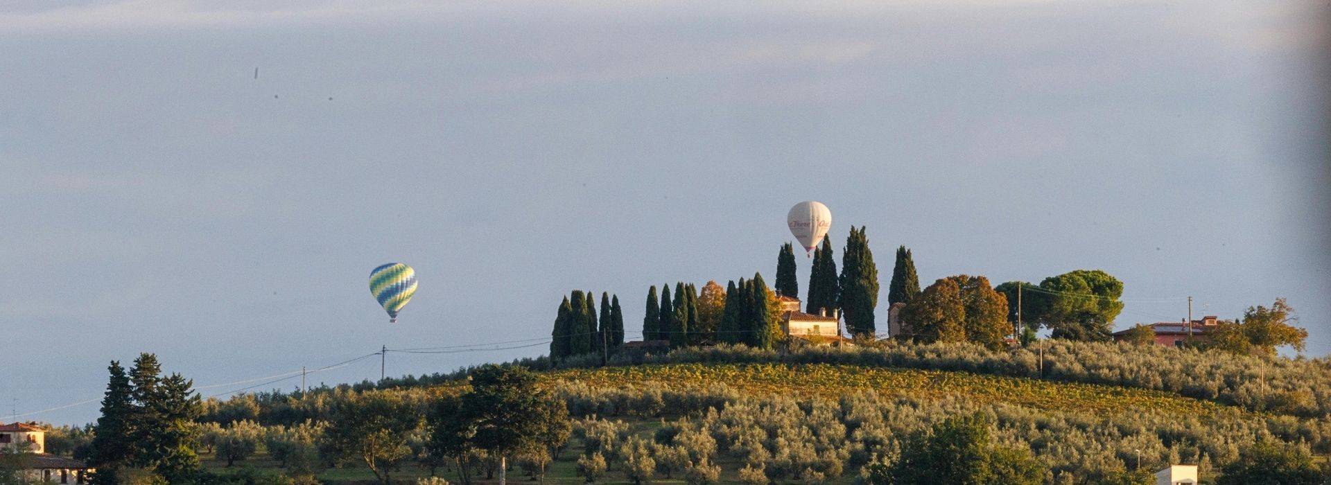 Two hot air balloons flying at dawn over a Tuscan hill with vineyards, olive trees, cypresses, and a rural farmhouse nestled in the Chianti countryside