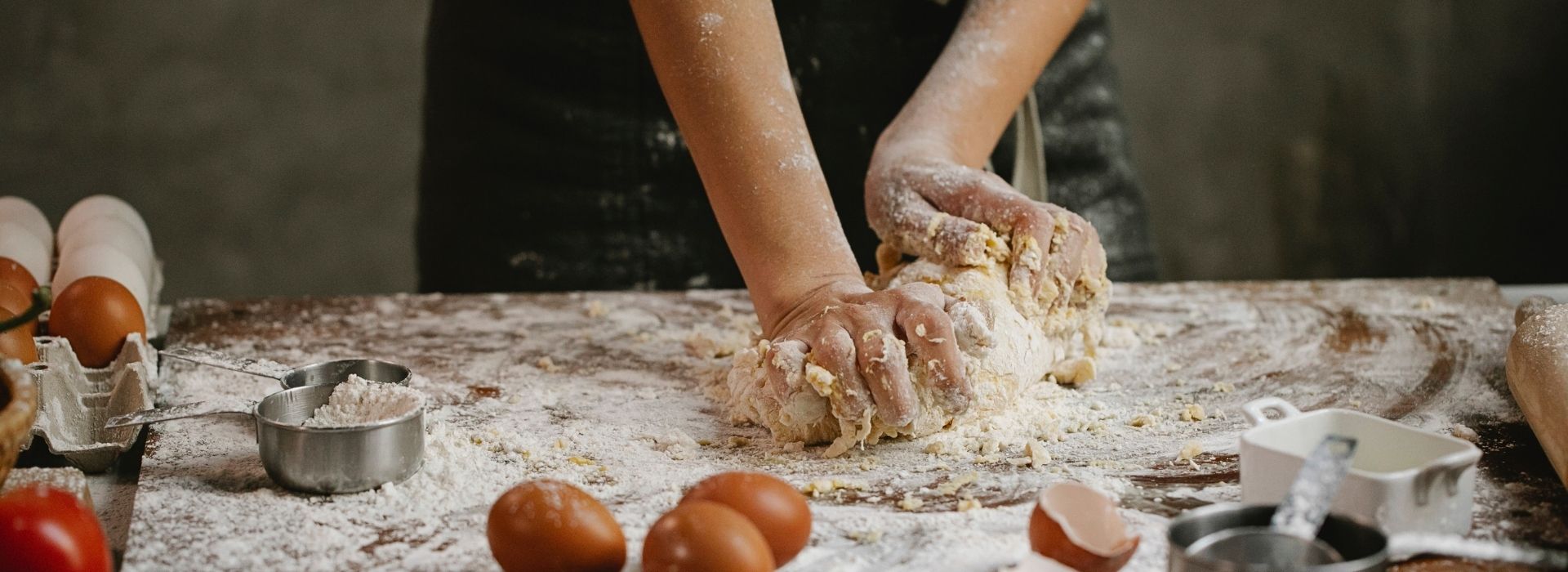 Flourished hands kneading fresh pasta on a floured wooden table, with eggs, eggshells, and a measuring cup in the foreground during a cooking class in Maremma