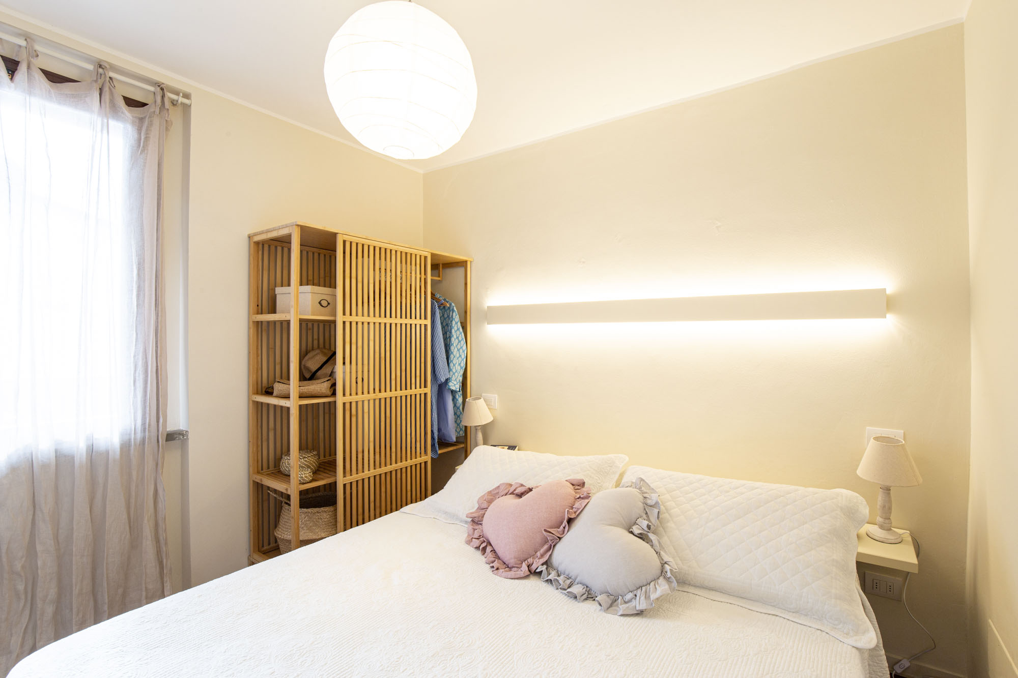 Panoramic view of the interior of the double bedroom in the Terrace View Studio. In the foreground is the bed with a white quilted coverlet and two heart-shaped pillows (pink and grey) with ruffle trim. On the left wall, an open bamboo wardrobe features shelves and hanging garments. Above the headboard, a modern linear LED sconce diffuses a warm, soft light, while a spherical rice paper pendant lamp hangs from the ceiling. On the right, a white nightstand with a coordinating lamp and the window with a light grey linen curtain. The atmosphere is cozy and relaxing.