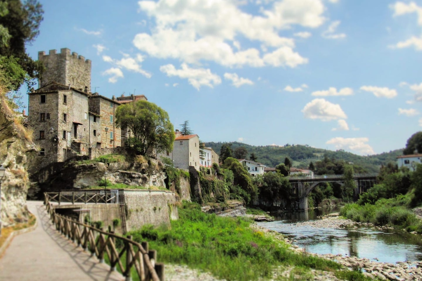 Medieval tower of Subbiano Castle standing on a rocky crag above the Arno River, with a promenade along the bank, an arched bridge in the background and green Tuscan hills under a blue sky