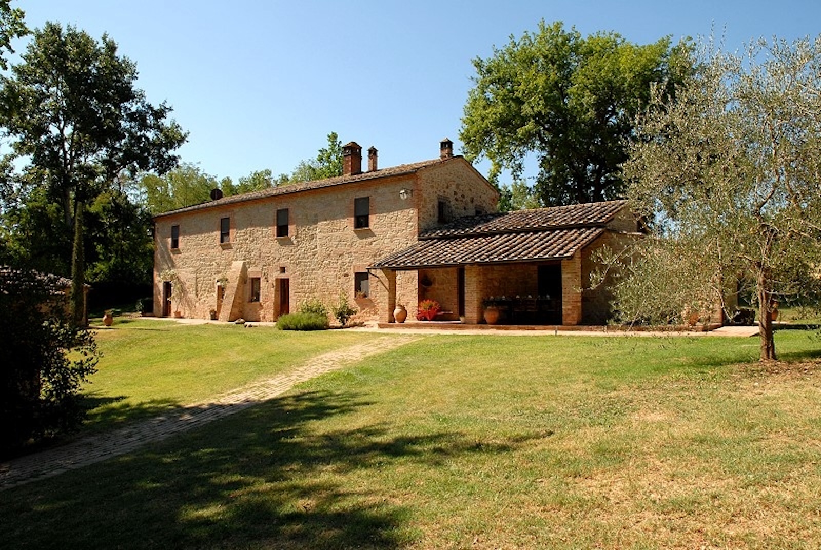 Exterior view of a beautifully restored historic stone Tuscan farmhouse, set within a spacious, well-kept green lawn. On the right, a covered brick porch features dining seating, with an olive tree in the foreground. A brick pathway crosses the lawn leading to the main property, which is surrounded by mature trees under a clear blue sky.