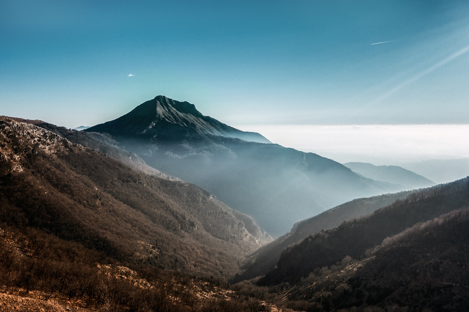Veduta panoramica del Monte Matanna nelle Alpi Apuane meridionali, con la cima che si staglia contro un cielo azzurro e vallate ricoperte di boschi avvolte nella foschia autunnale