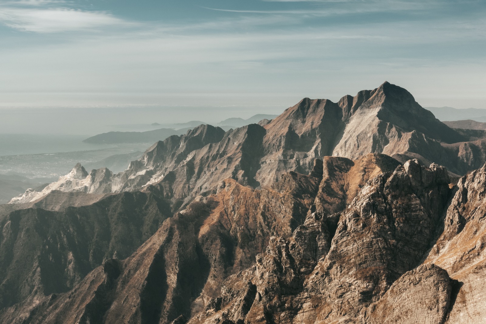 Veduta aerea e panoramica delle vette rocciose delle Alpi Apuane, con i versanti frastagliati, cave di marmo visibili lungo i pendii e la costa con il mare all'orizzonte sotto un cielo limpido