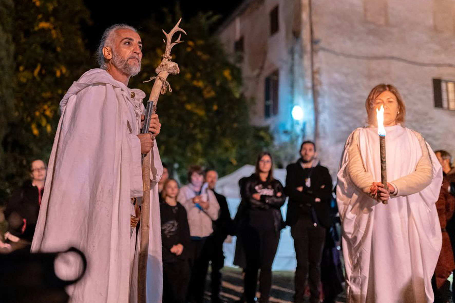 Personaggio in costume con bastone durante un evento serale nel centro storico, con il pubblico sullo sfondo.