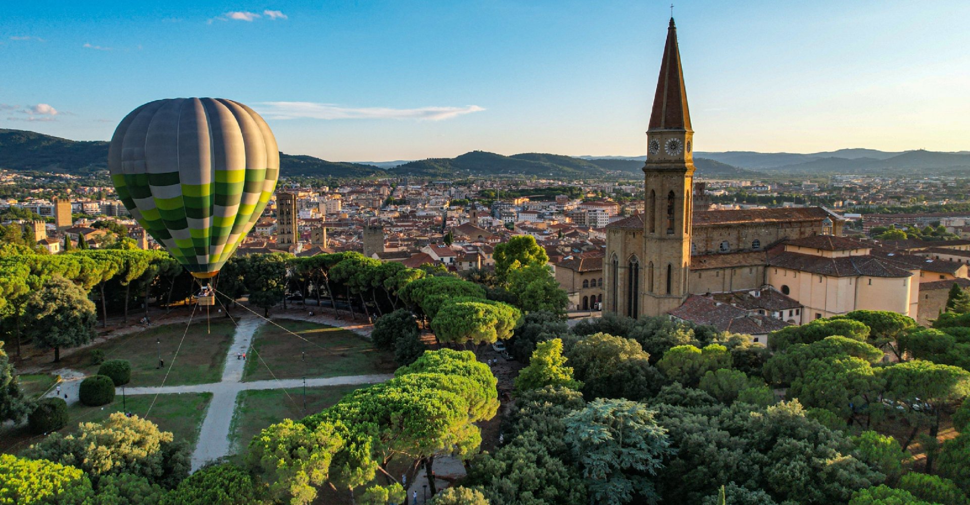 Aerial view of Arezzo: in the foreground is the lush Parco del Prato, with crisscrossing paths and a large hot-air balloon in flight, flanked on the right by the imposing Duomo and its tall bell tower.
