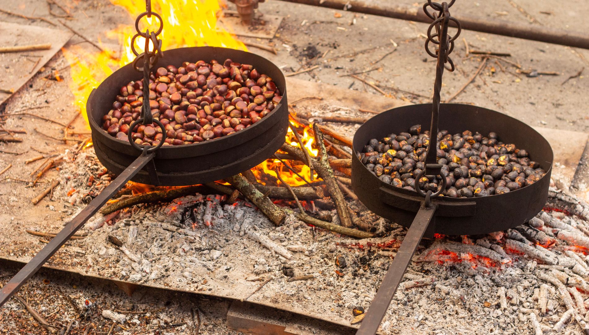 Castagne in cottura in due grandi padelle sul fuoco di legna, tra fiamme e braci.