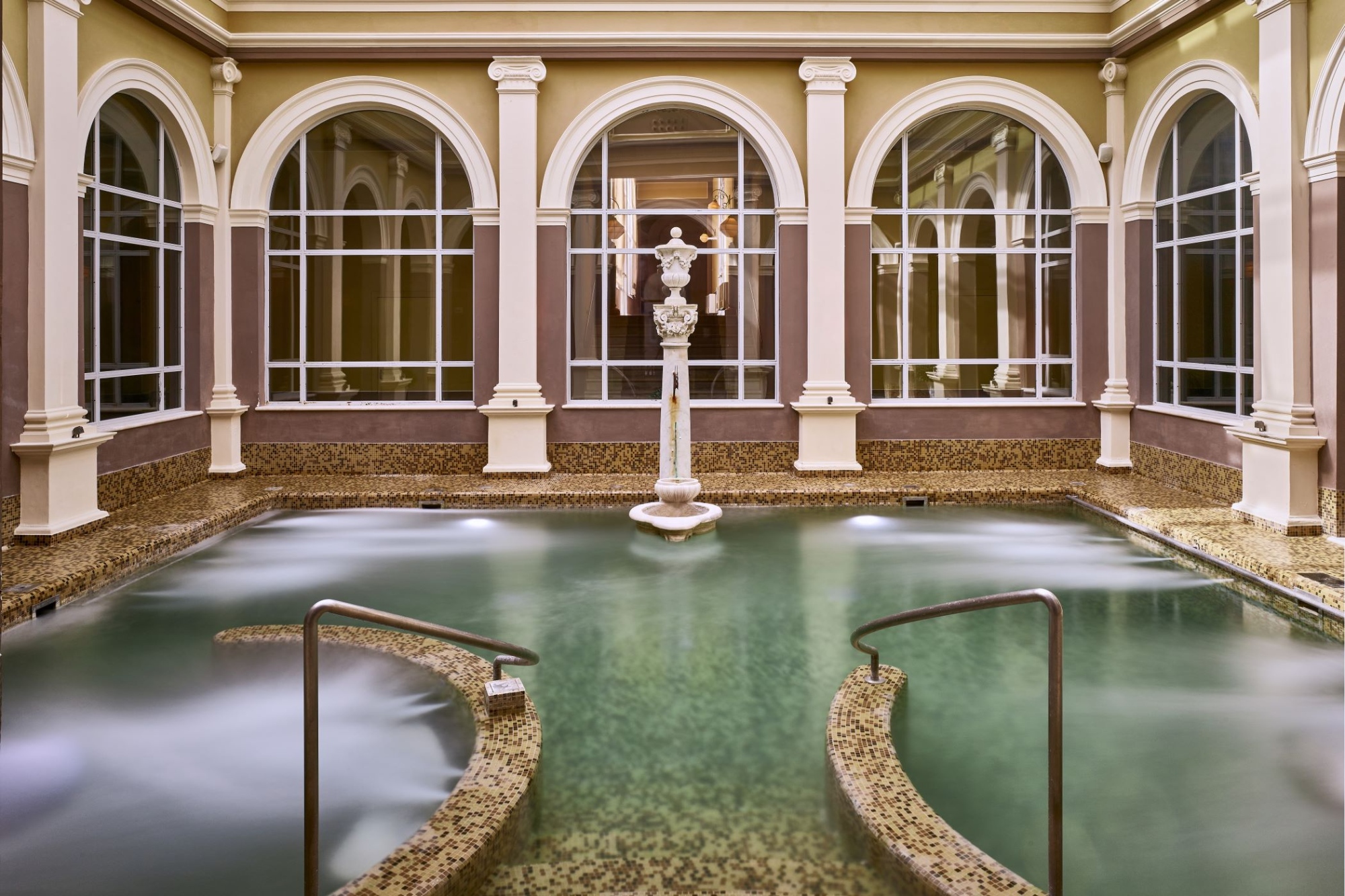 Piscina termale coperta in stile classico con acqua verde, colonnati bianchi, grandi finestre ad arco, vasca di marmo con scalini e statue decorative, zona benessere dell'elegante struttura termale
