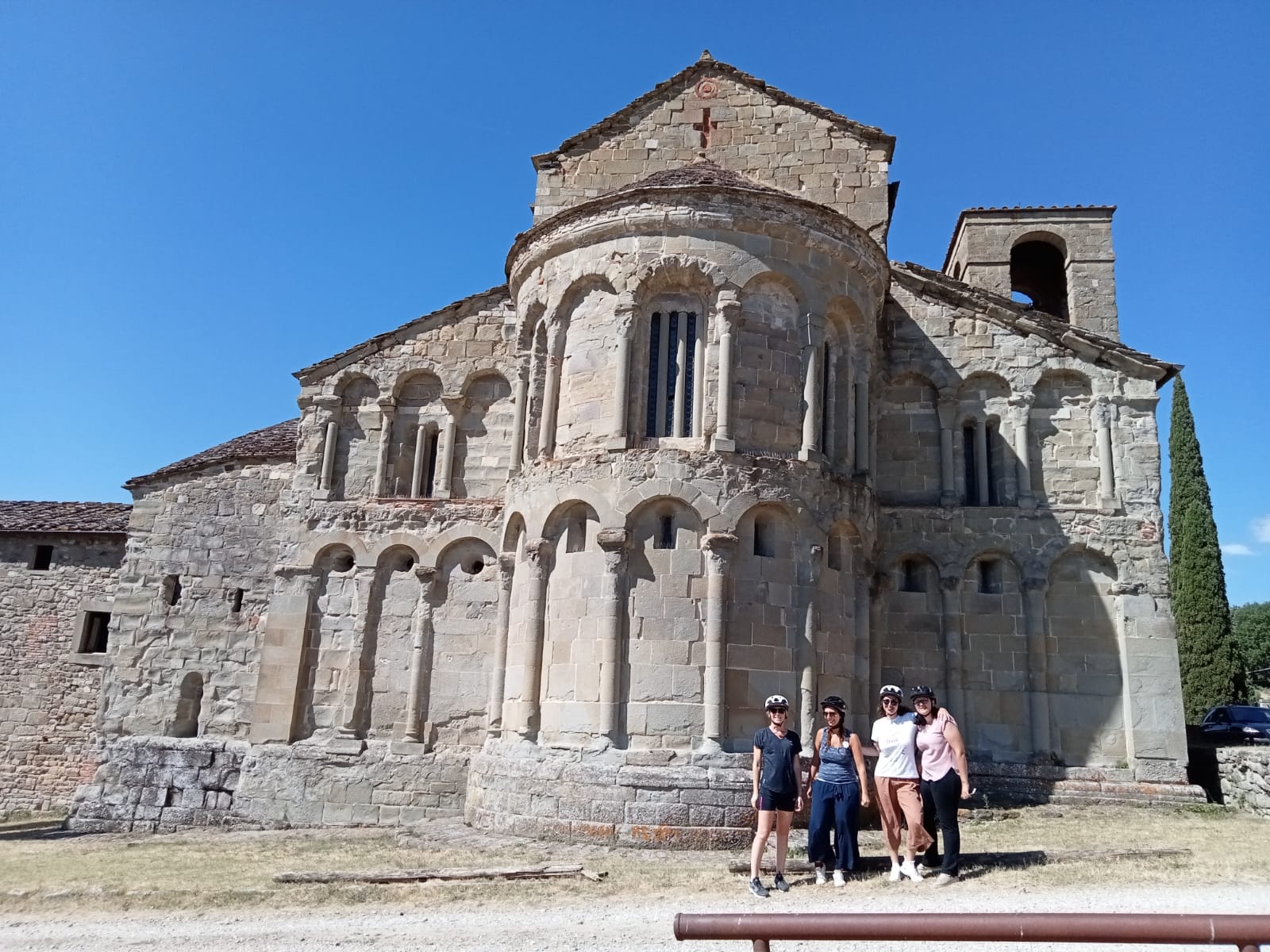 Quattro cicliste con casco in posa davanti all'abside romanica della Pieve di Romena, edificio in pietra arenaria con arcate cieche e campanile, in una giornata di sole nel Casentino