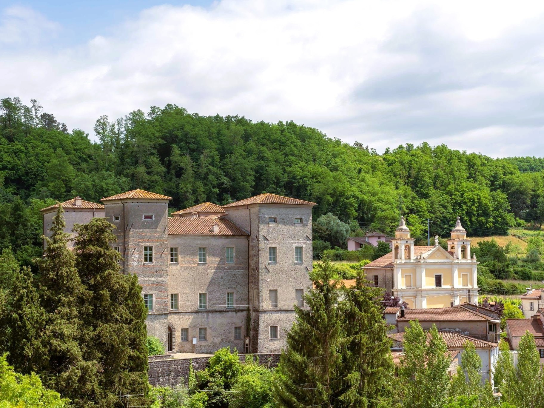 Vista panoramica del borgo di Pontebosio con il Castello dei Malaspina in primo piano, la chiesa barocca gialla con campanile e il bosco verde sullo sfondo, nella Valle del Taverone in Lunigiana