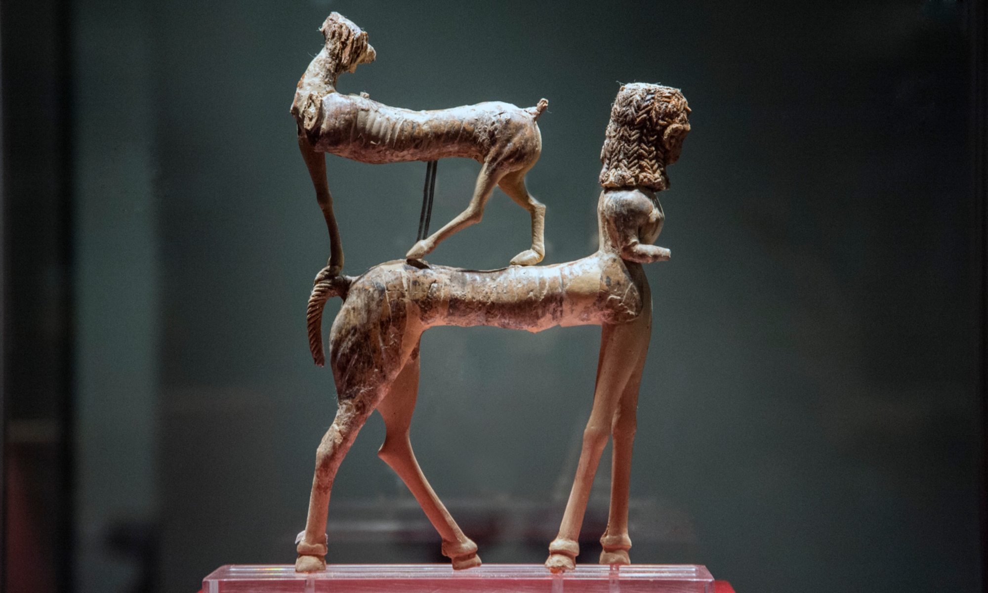 Statuetta etrusca in bronzo con due figure animali sovrapposte, esposta su supporto trasparente