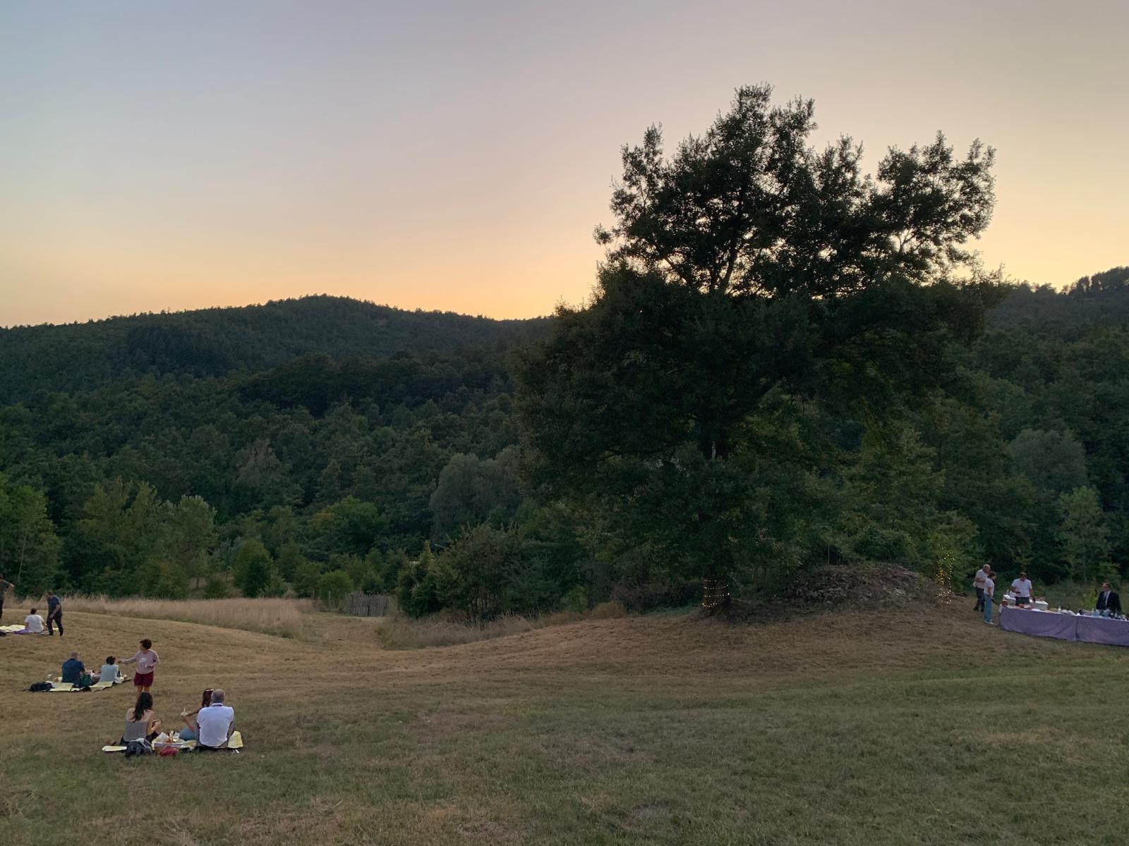 Tramonto sulle colline boscose di Chiusi della Verna con persone sedute su coperte da picnic in un prato durante la Cena Stellata a Quercus La Baita