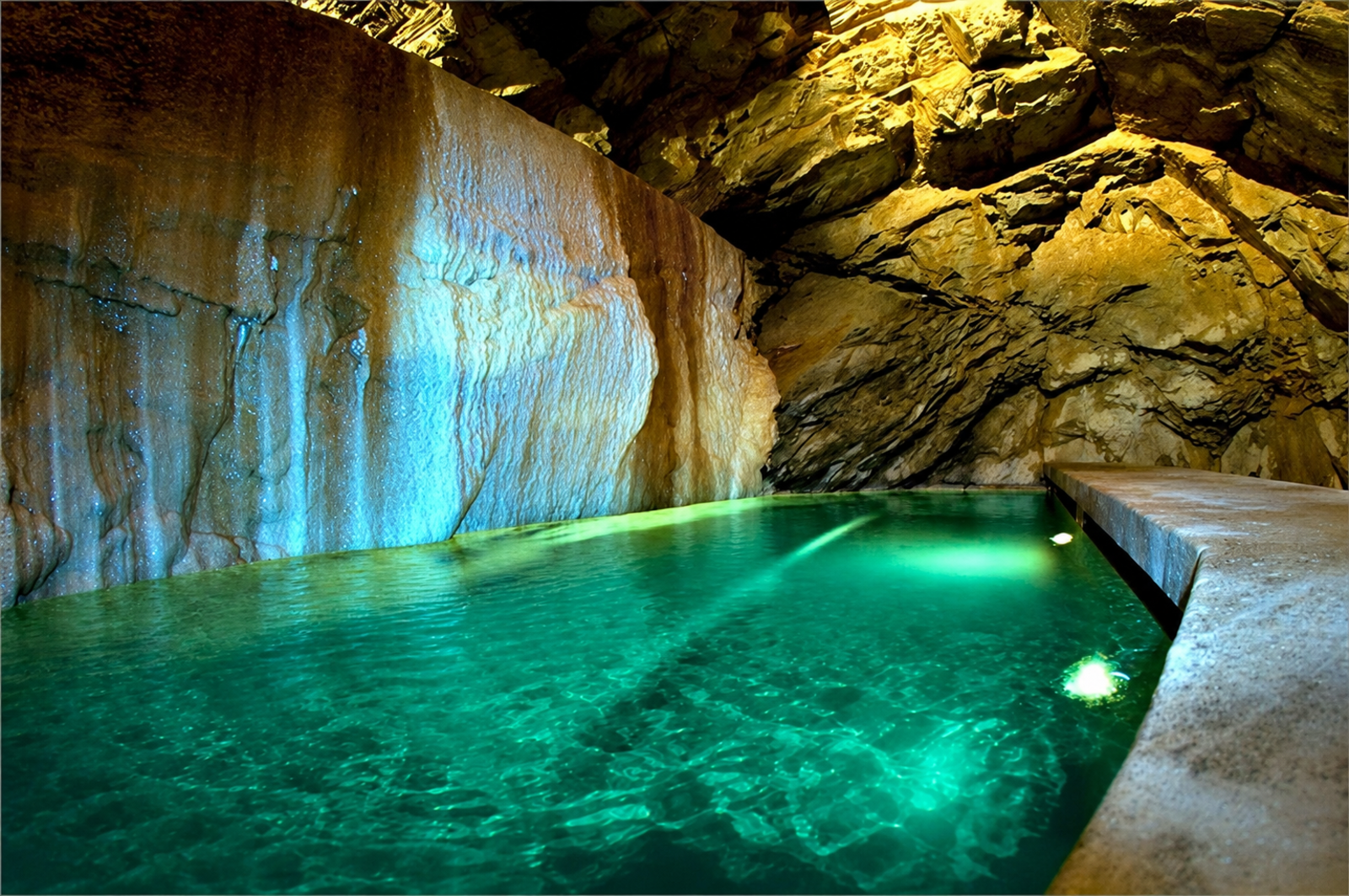 Grotta termale sotterranea con piscina di acqua termale verde smeraldo illuminata artificialmente, pareti di roccia gialla e piattaforma in cemento per l'accesso all'interno delle Terme di Bagni di Pisa