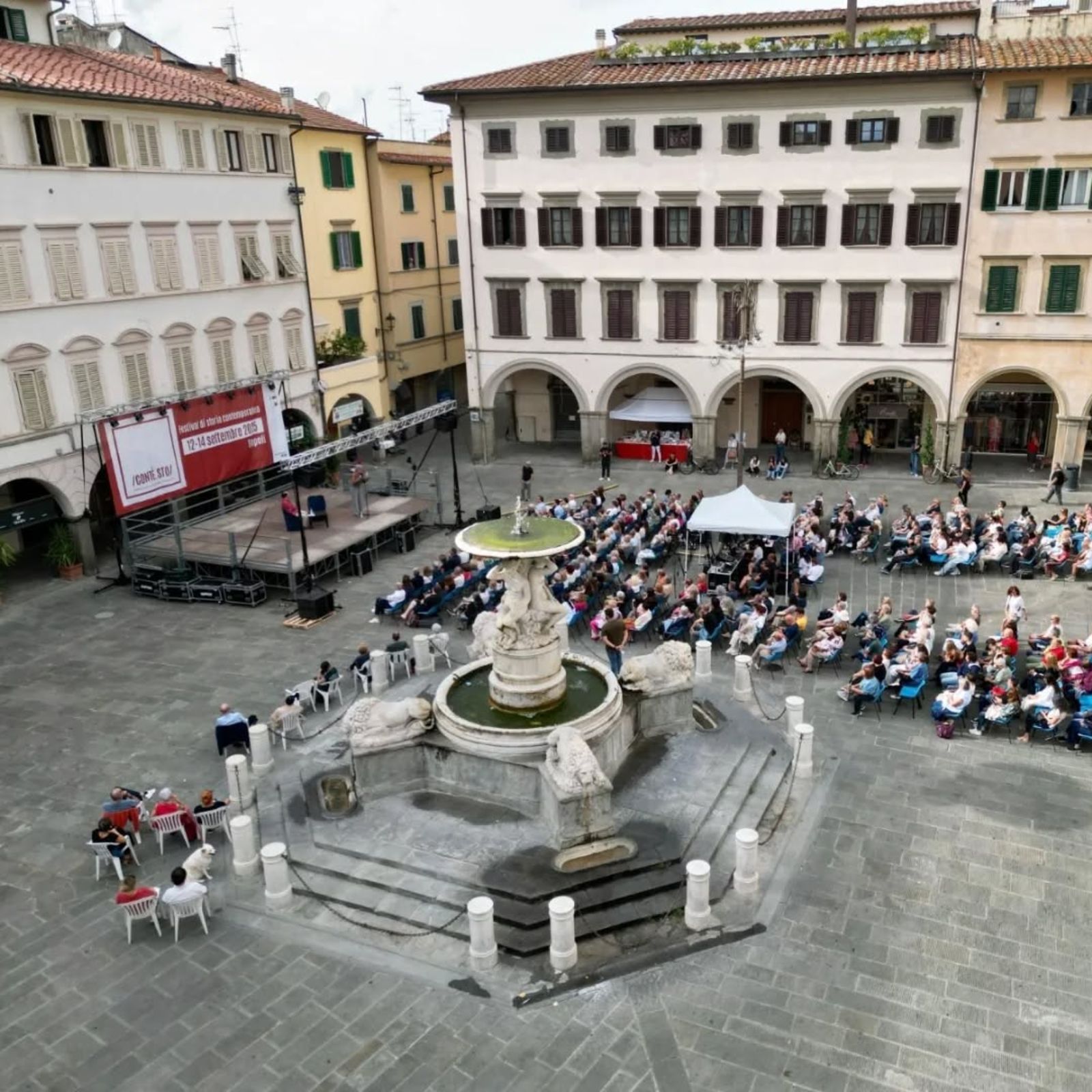 Piazza con fontana monumentale al centro e pubblico seduto per un evento all’aperto, davanti a un palco e agli edifici storici.