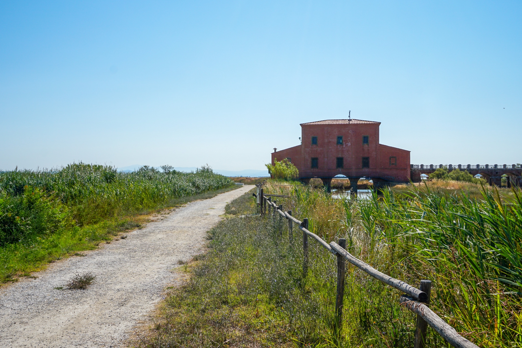 Im Vordergrund ein Feldweg, der an einem Sumpfgebiet entlangführt und zur Casa Rossa Ximenes führt, einem historischen, rot verputzten Gebäude, das Bögen im Erdgeschoss prägen