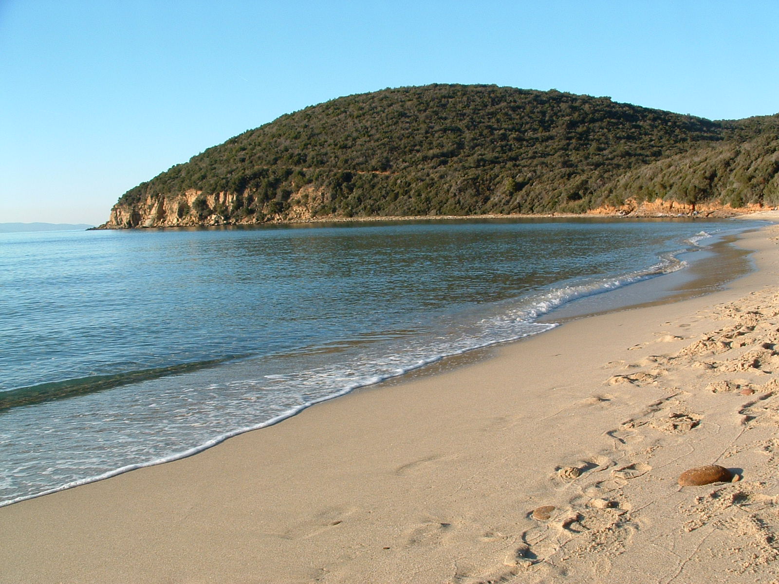 Spiaggia sabbiosa deserta lungo la costa della Maremma tra Punta Ala e le Rocchette, con onde leggere che lambiscono la riva, un promontorio verde ricoperto dalla macchia mediterranea a picco sul mare e cielo azzurro limpido al tramonto