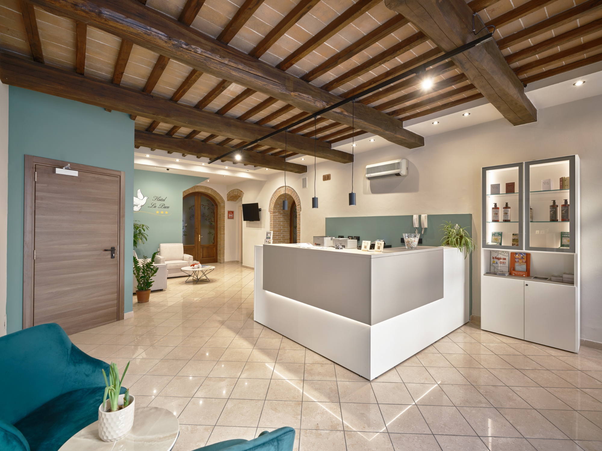 a reception moderna ed accogliente di un hotel, che unisce elementi rustici come un soffitto a travi in legno a vista a un arredamento contemporaneo. Lo spazio è caratterizzato da pareti chiare con accenti verde salvia e un pavimento lucido in piastrelle. Al centro domina un grande bancone reception minimalista, bianco e grigio tortora, illuminato da LED. Sulla destra è presente un mobile espositore con prodotti in vendita, mentre sulla sinistra si trova una zona lounge con poltroncine in velluto e un tavolino. Sullo sfondo si legge il logo 