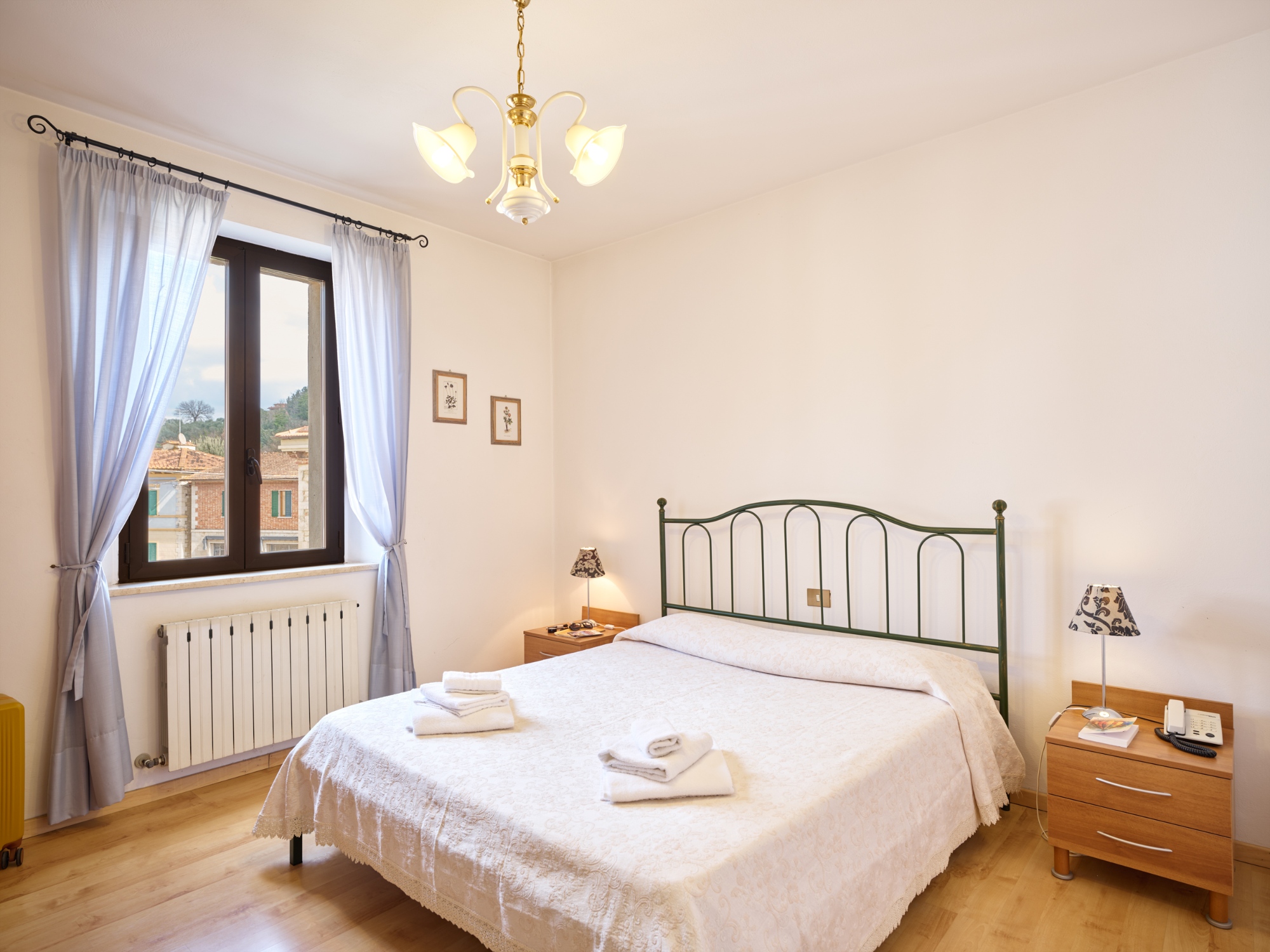 Tuscan-style double bedroom featuring a green wrought iron bed, parquet flooring, and a panoramic view of a historic village and vineyards through the window.