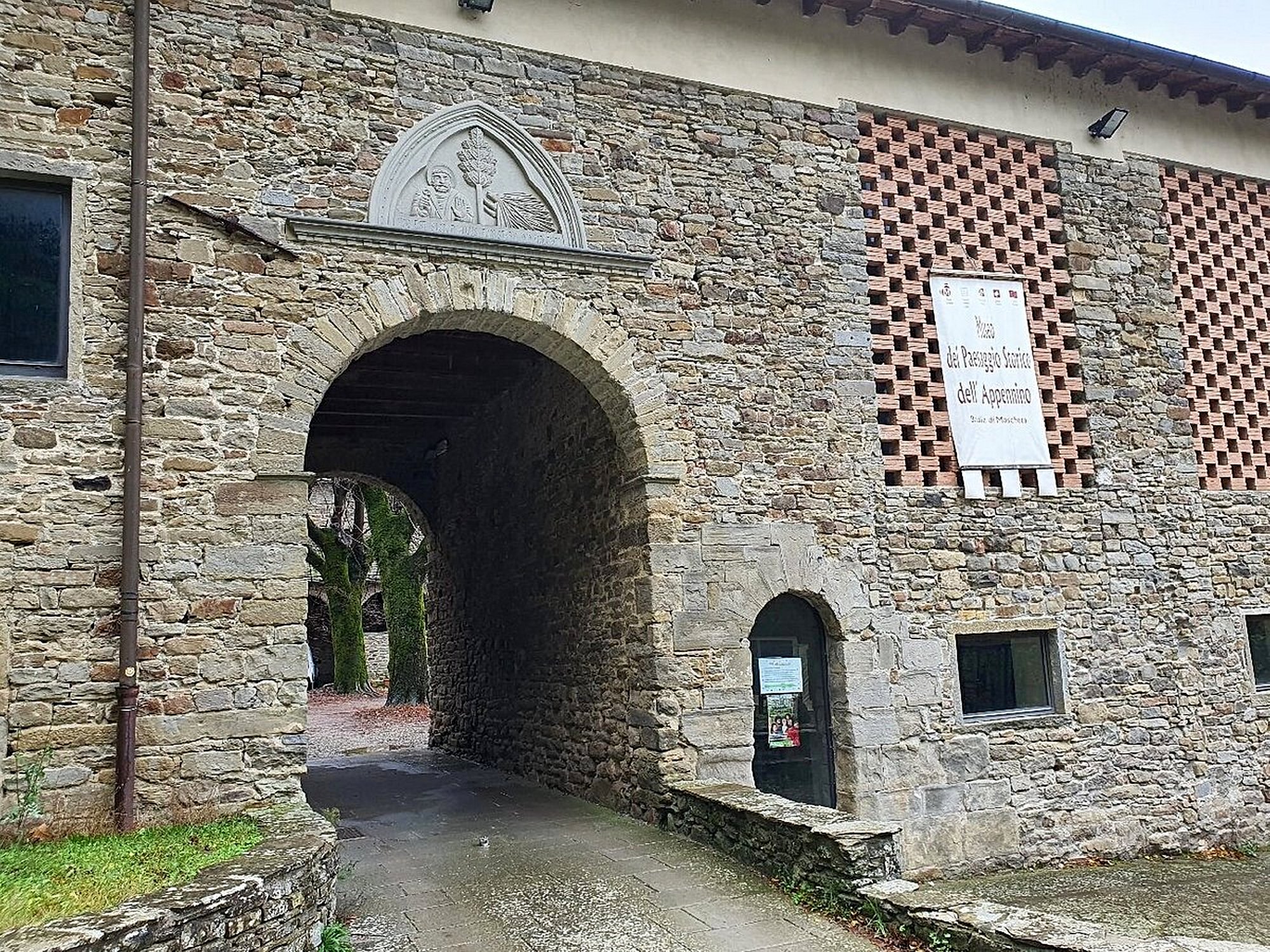 Dettaglio architettonico dell'ingresso della Badia di Moscheta con grande arco in pietra, lunetta decorata e finestra rettangolare con pattern geometrico in mattoni rossi e bianchi