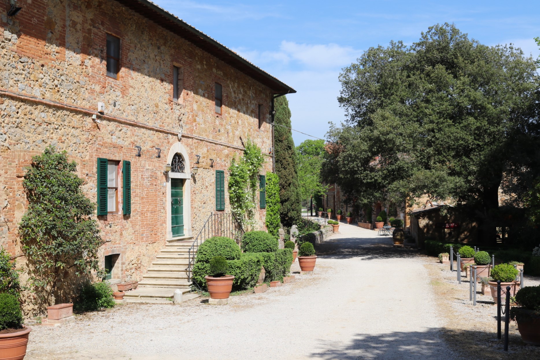The entrance to the farmhouse with a dirt road and a small building on the left surrounded by trees and plants