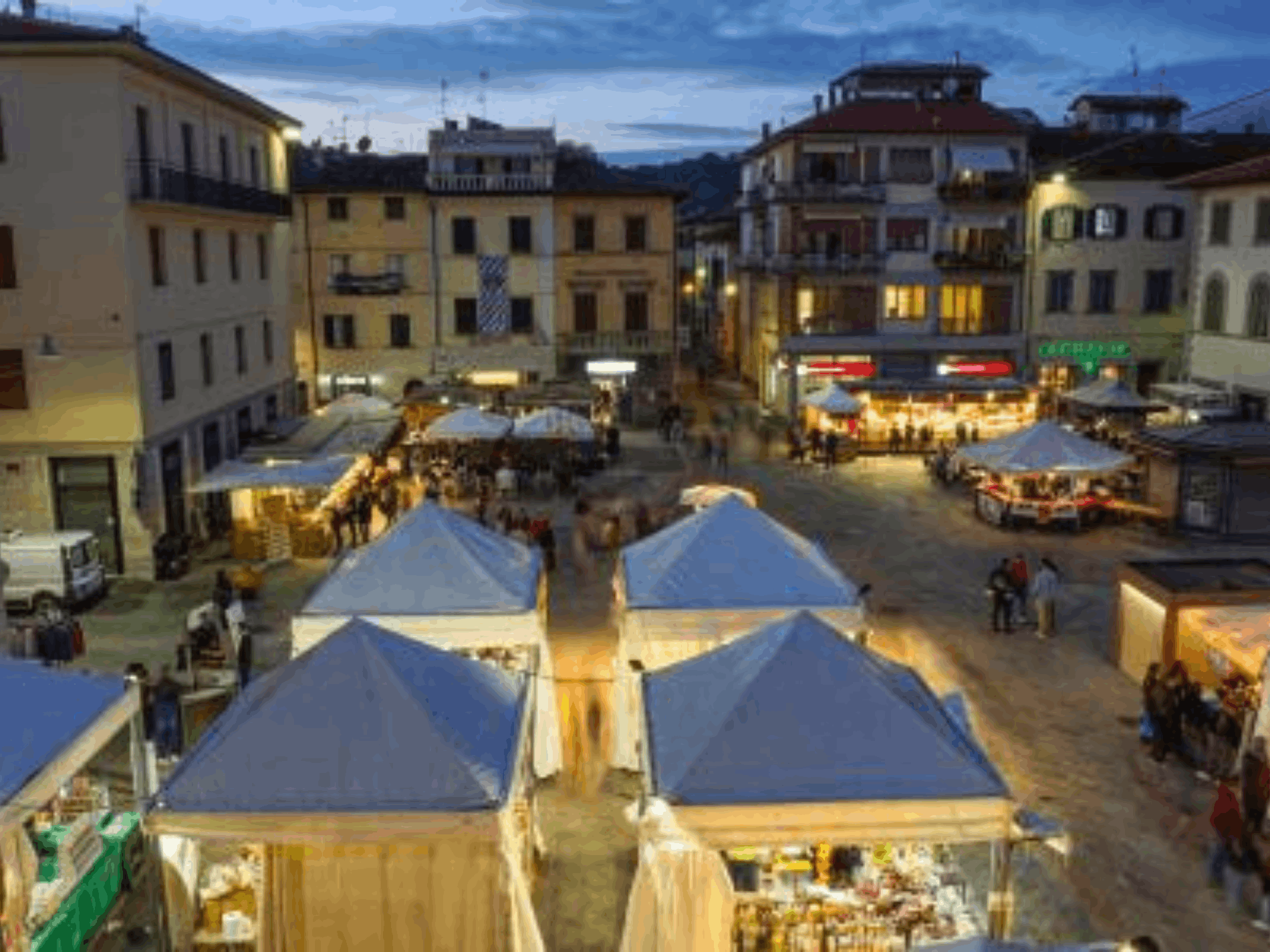 Piazza della Repubblica a Terranuova Bracciolini durante la Festa del Perdono al crepuscolo, con numerosi banchi di mercato illuminati e visitatori.